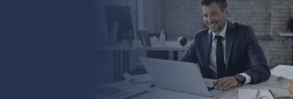 banner home - homem de terno sorrindo, em mesa de trabalho olhando para a tela do laptop
